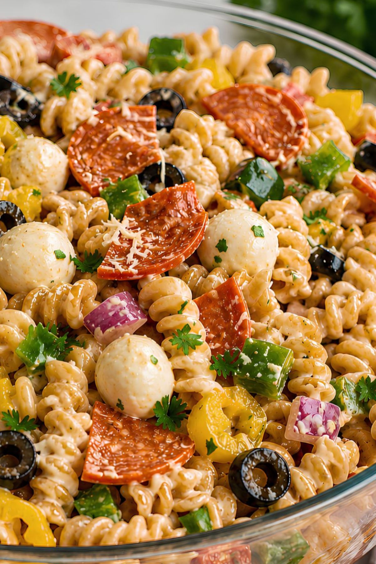High protein pizza pasta salad with creamy rotini, quartered turkey pepperoni, mozzarella pearls, black olives, green bell pepper, banana peppers, red onion, Parmesan, and fresh parsley in a clear glass bowl.
