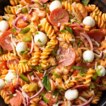 Close-up of high protein pizza pasta salad with rotini, pepperoni, mozzarella pearls, tomatoes, green bell peppers, red onion, and fresh herbs in a black bowl.