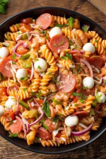 Close-up of high protein pizza pasta salad with rotini, pepperoni, mozzarella pearls, tomatoes, green bell peppers, red onion, and fresh herbs in a black bowl.