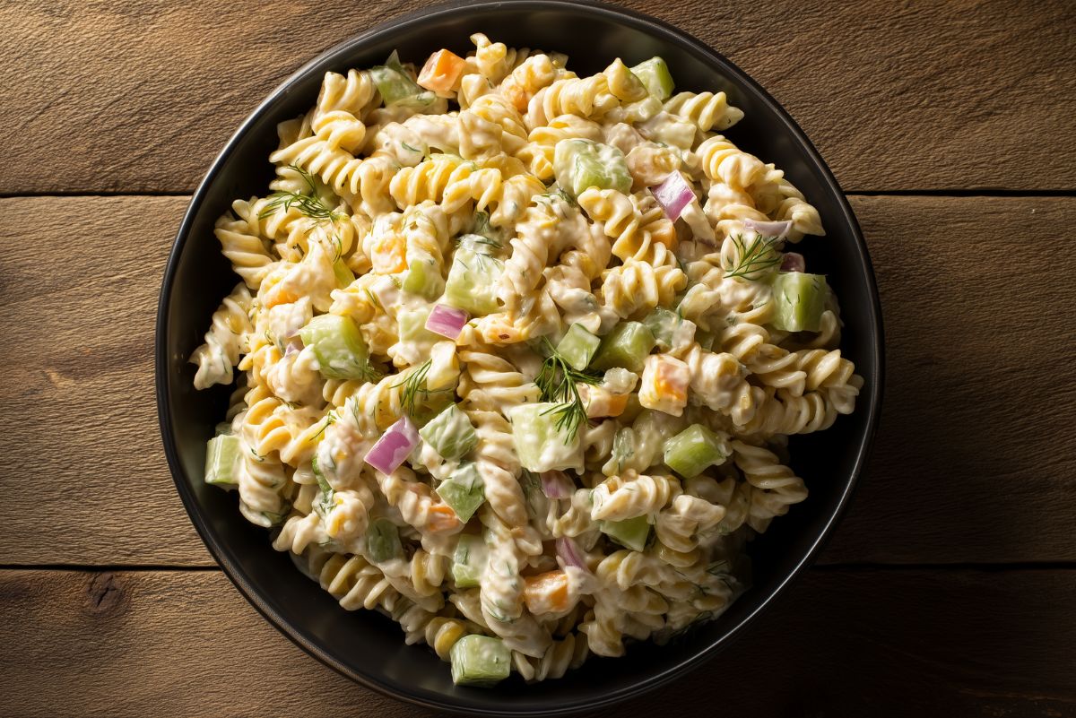 Overhead view of light dill pickle rotini pasta salad in a black bowl with creamy dressing, chopped pickles, red onion, cheddar cheese, and fresh dill on a rustic wood table.