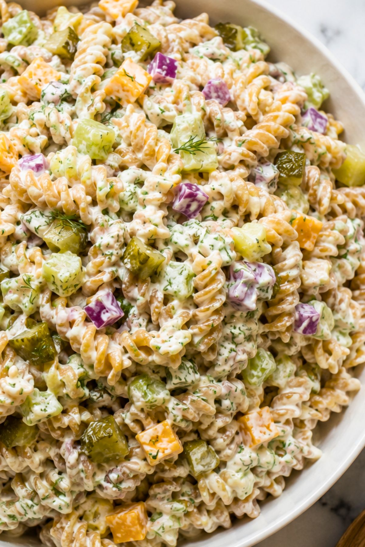 Creamy dill pickle rotini pasta salad with chopped pickles, cucumber, cheddar cheese, red onion, and fresh dill in a white serving bowl.