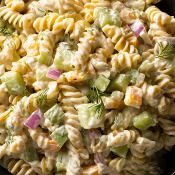 Close-up of creamy light dill pickle rotini pasta salad with chopped pickles, red onion, cheddar cheese, and fresh dill in a black bowl.