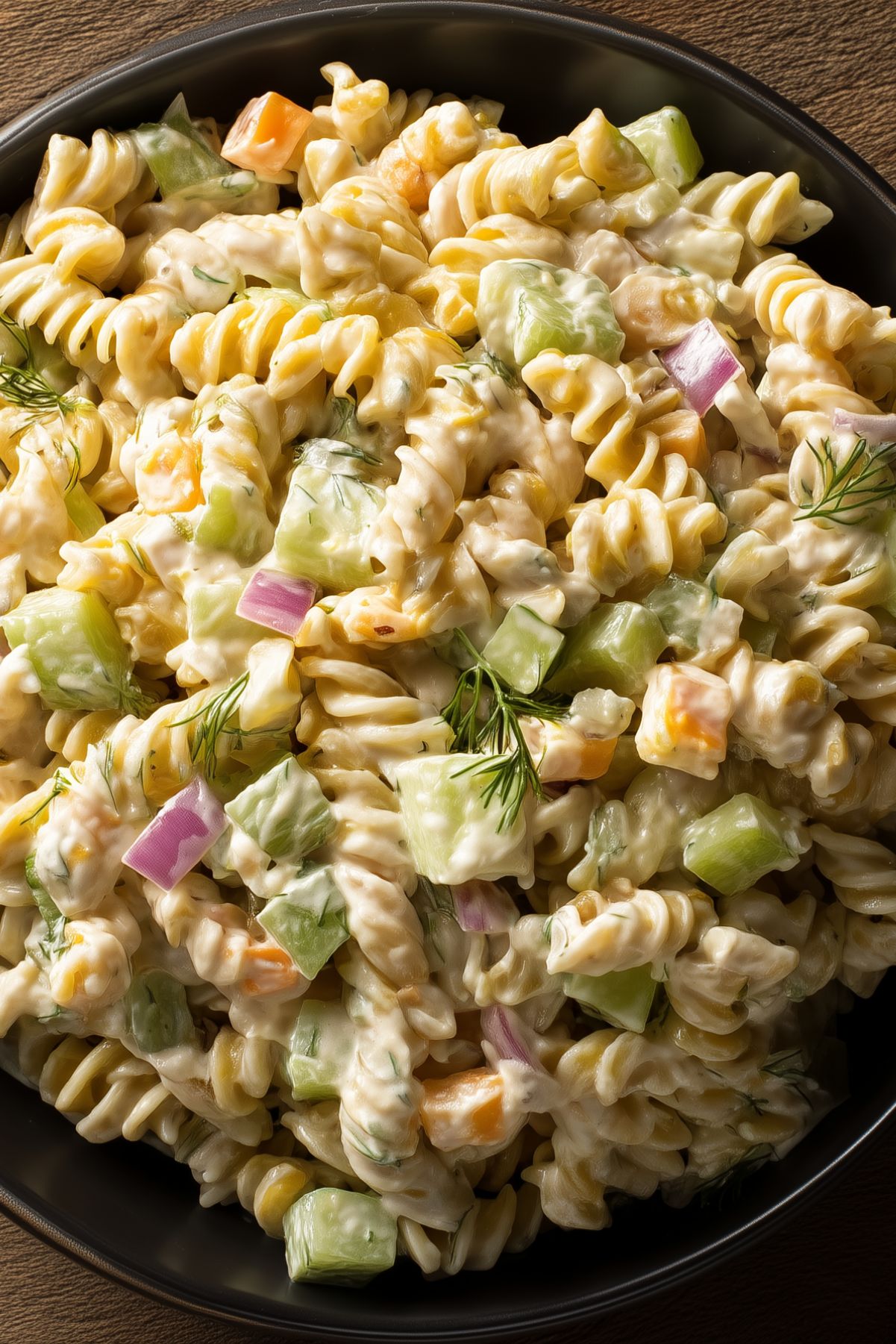 Close-up of creamy light dill pickle rotini pasta salad with chopped pickles, red onion, cheddar cheese, and fresh dill in a black bowl.