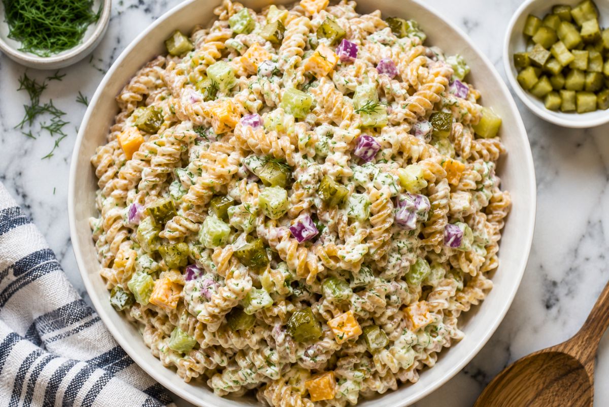 Overhead view of creamy dill pickle rotini pasta salad with chopped pickles, cucumber, cheddar cheese, red onion, and fresh dill, served beside extra pickles and herbs.