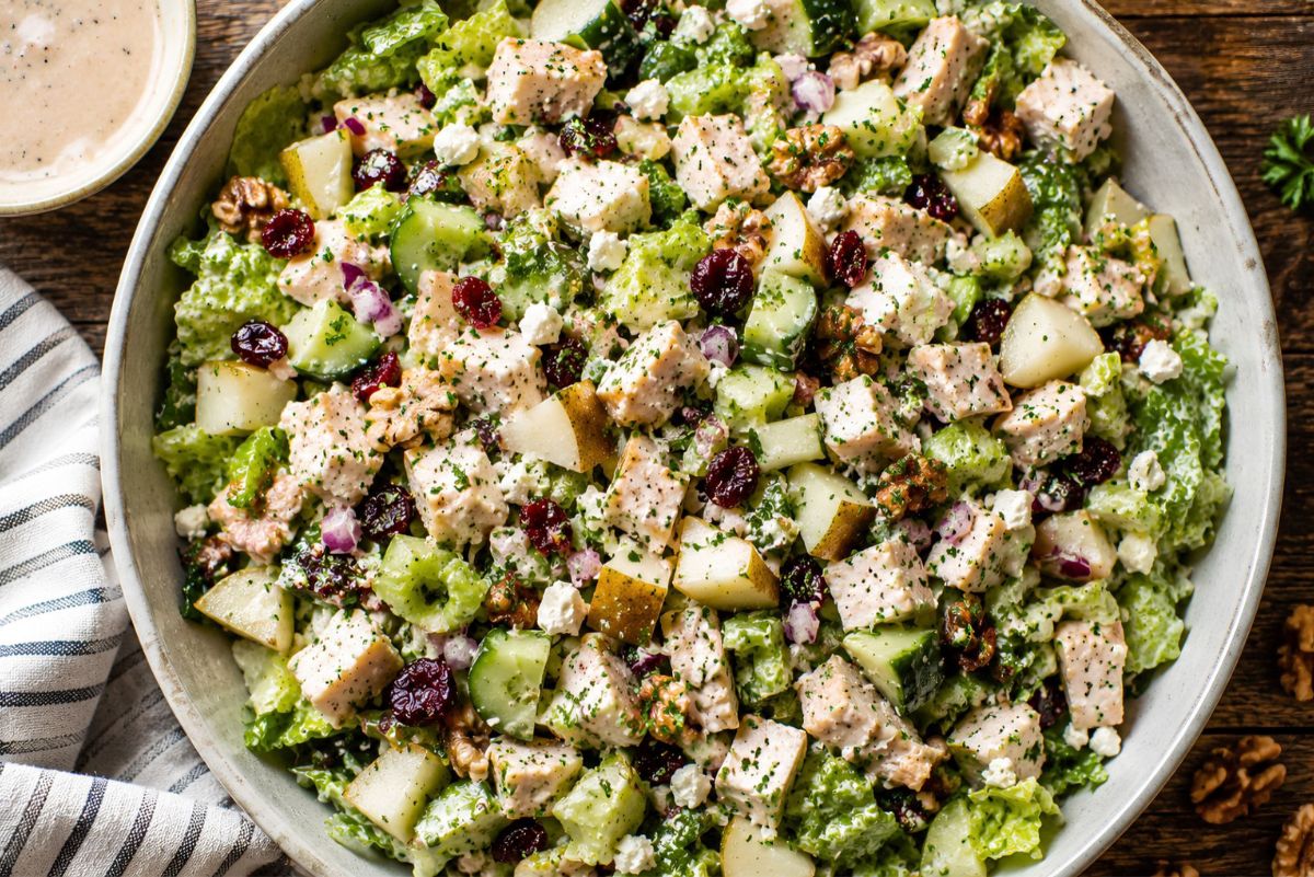 Overhead bowl of pear walnut chicken chopped salad with romaine, cucumber, celery, diced pear, dried cranberries, red onion, feta, walnuts, and chicken in creamy balsamic Dijon dressing, served on a rustic wooden table.