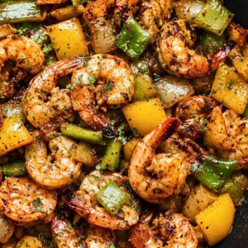 Close-up of Cajun shrimp sautéed with chunky yellow and green peppers and onions in a dark skillet, finished with chopped parsley and black pepper.