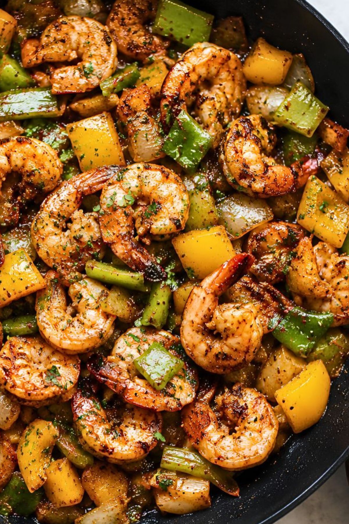 Close-up of Cajun shrimp sautéed with chunky yellow and green peppers and onions in a dark skillet, finished with chopped parsley and black pepper.