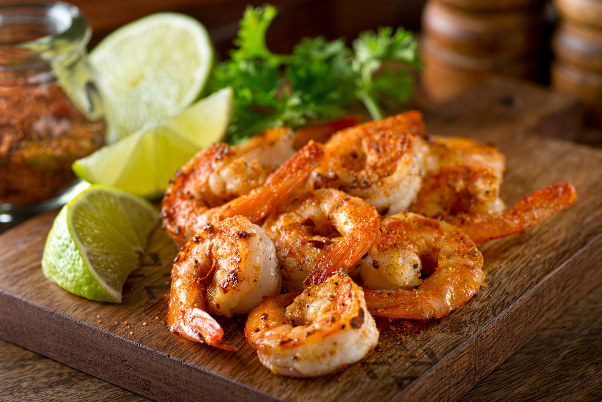 Cajun-seasoned shrimp arranged on a wooden cutting board with lime wedges, lemon, parsley, and a jar of spices in the background.