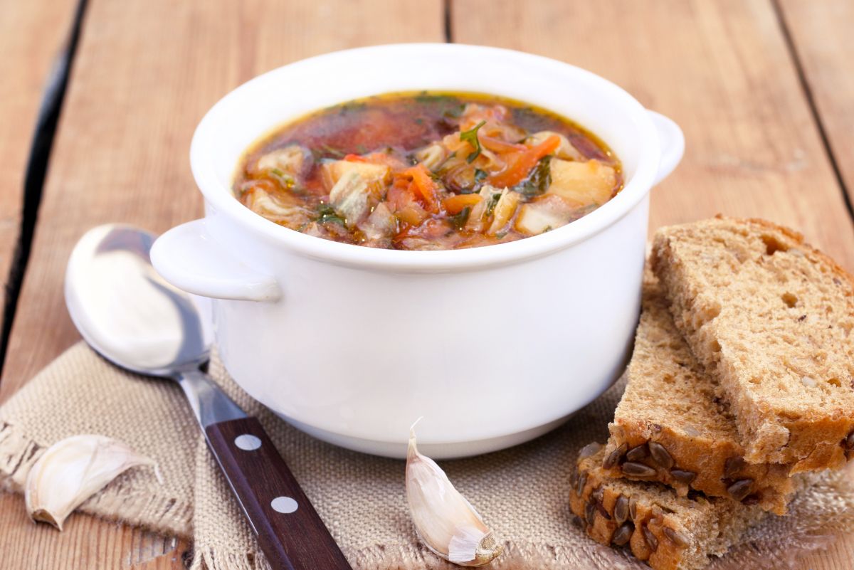 Rustic chicken cabbage and tomato soup served in a white bowl, with visible chicken, cabbage, carrots, tomatoes, and herbs. Slices of seeded bread, garlic cloves, and a spoon rest beside the bowl on a wooden table.