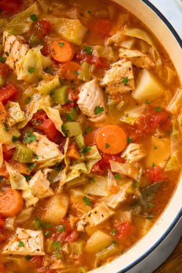 Close-up of rustic chicken cabbage and tomato soup with tender chicken, chopped cabbage, carrots, celery, potatoes, diced tomatoes, parsley, and a bay leaf in a rich tomato broth.