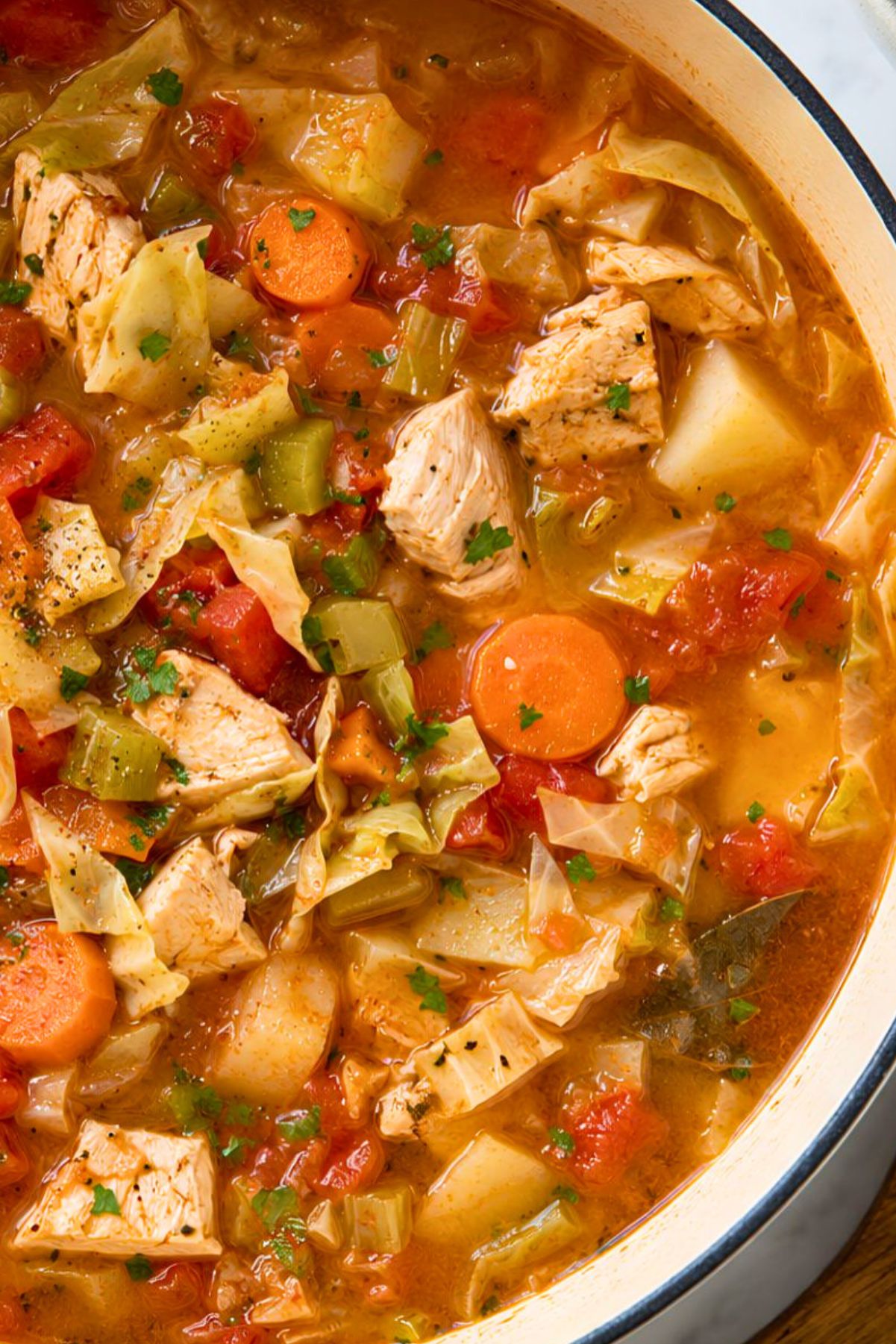 Close-up of rustic chicken cabbage and tomato soup with tender chicken, chopped cabbage, carrots, celery, potatoes, diced tomatoes, parsley, and a bay leaf in a rich tomato broth.