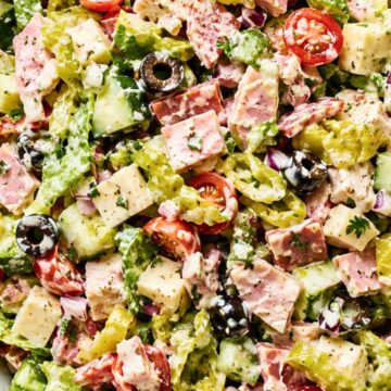 Spicy Italian Grinder Chopped Salad with chopped romaine, deli meats, provolone cheese, cherry tomatoes, cucumbers, banana peppers, black olives, red onion, herbs, and creamy grinder dressing in a large bowl.