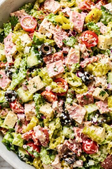 Spicy Italian Grinder Chopped Salad with chopped romaine, deli meats, provolone cheese, cherry tomatoes, cucumbers, banana peppers, black olives, red onion, herbs, and creamy grinder dressing in a large bowl.