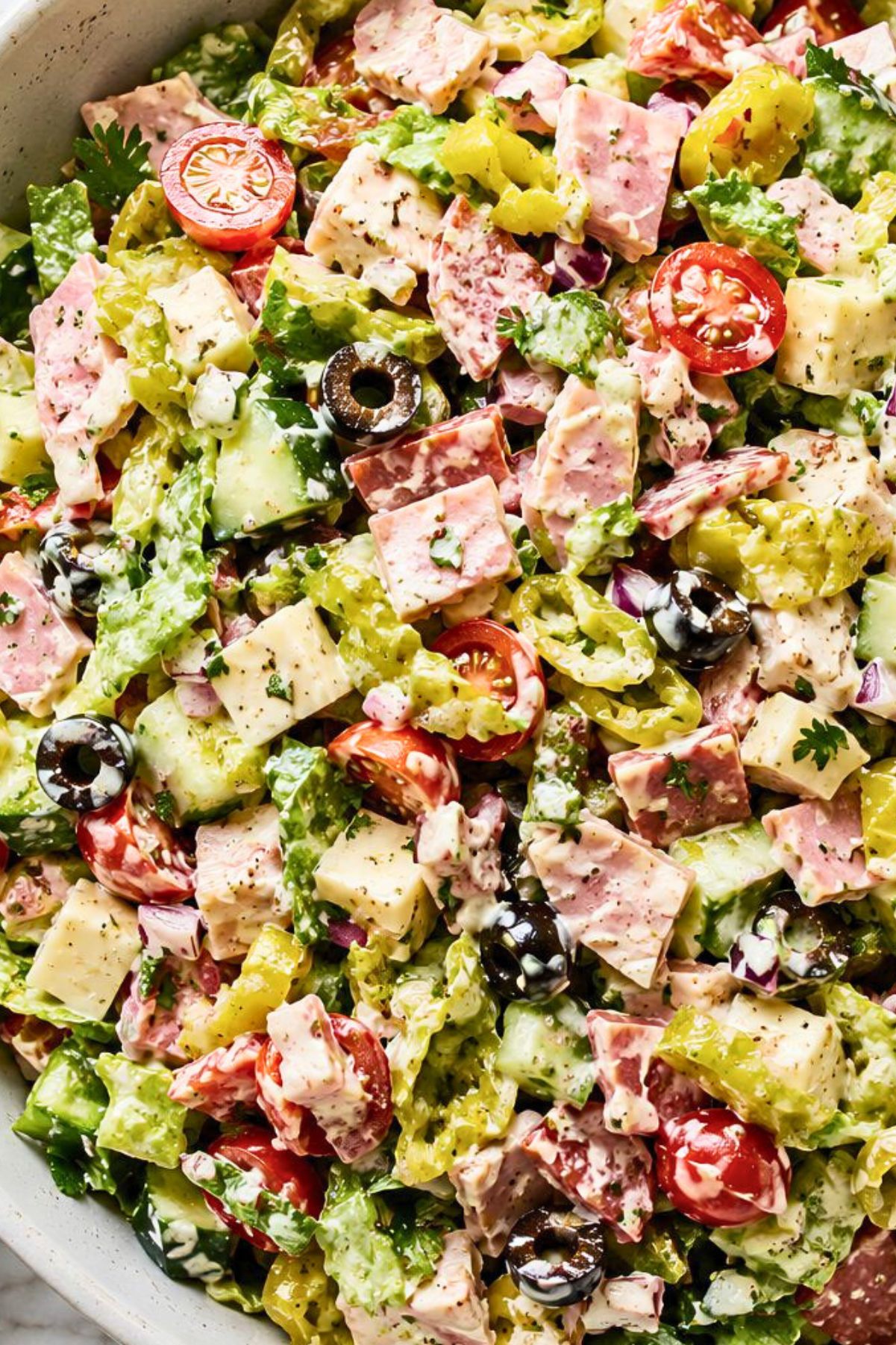 Spicy Italian Grinder Chopped Salad with chopped romaine, deli meats, provolone cheese, cherry tomatoes, cucumbers, banana peppers, black olives, red onion, herbs, and creamy grinder dressing in a large bowl.