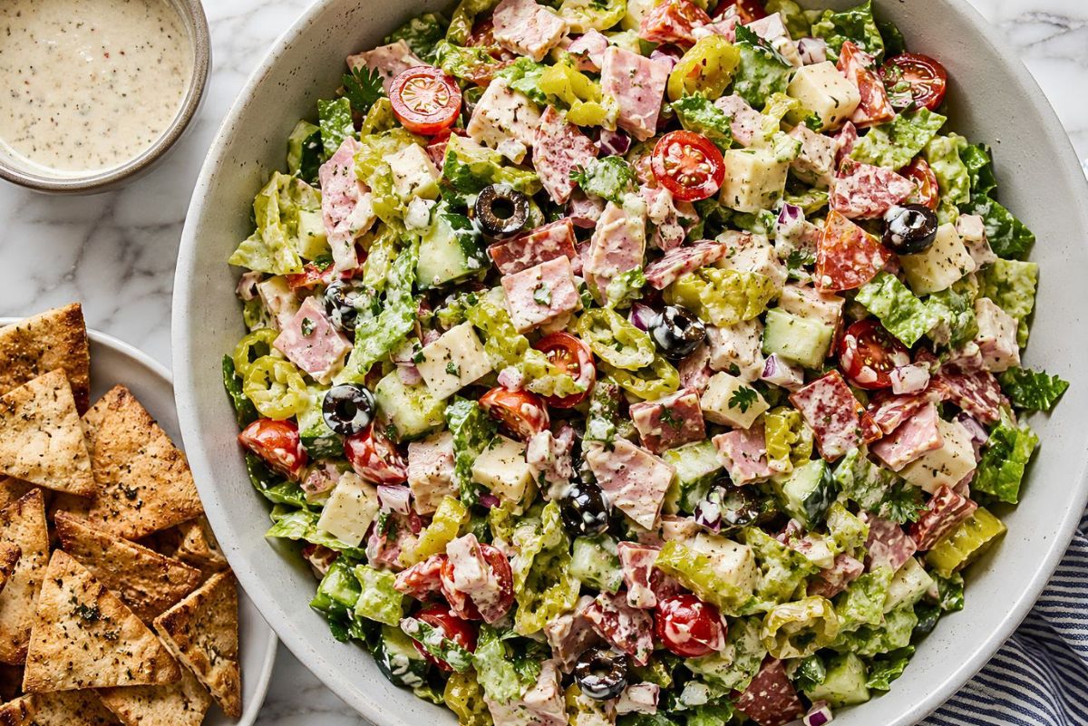 Spicy Italian Grinder Chopped Salad in a large bowl with romaine, deli meats, provolone, cherry tomatoes, cucumbers, banana peppers, black olives, red onion, and creamy grinder dressing, served with garlic pita chips on the side.