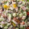Close-up of spicy Italian grinder chopped salad with salami, lettuce, cucumbers, tomatoes, black olives, red onion, peppers, and creamy Italian dressing in a black bowl.