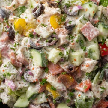 Close-up of spicy Italian grinder chopped salad with salami, lettuce, cucumbers, tomatoes, black olives, red onion, peppers, and creamy Italian dressing in a black bowl.