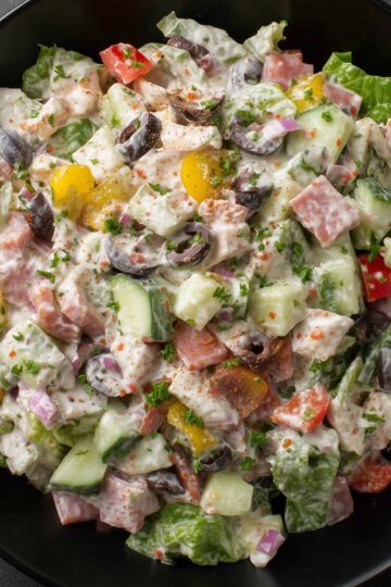 Close-up of spicy Italian grinder chopped salad with salami, lettuce, cucumbers, tomatoes, black olives, red onion, peppers, and creamy Italian dressing in a black bowl.