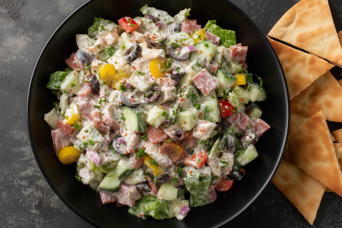 Overhead view of spicy Italian grinder chopped salad with lettuce, salami, cucumbers, tomatoes, black olives, red onion, peppers, and creamy dressing, served with pita chips.