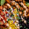Close-up of a Tex-Mex chicken sweet potato bowl with roasted sweet potatoes, seasoned chicken, black beans, corn, tomatoes, lettuce, shredded cheese, red onion, and creamy dressing.
