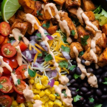 Close-up of a Tex-Mex chicken sweet potato bowl with roasted sweet potatoes, seasoned chicken, black beans, corn, tomatoes, lettuce, shredded cheese, red onion, and creamy dressing.