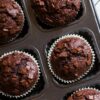 Chocolate oatmeal mini muffins topped with chocolate chunks in white paper liners and arranged in a dark muffin pan.