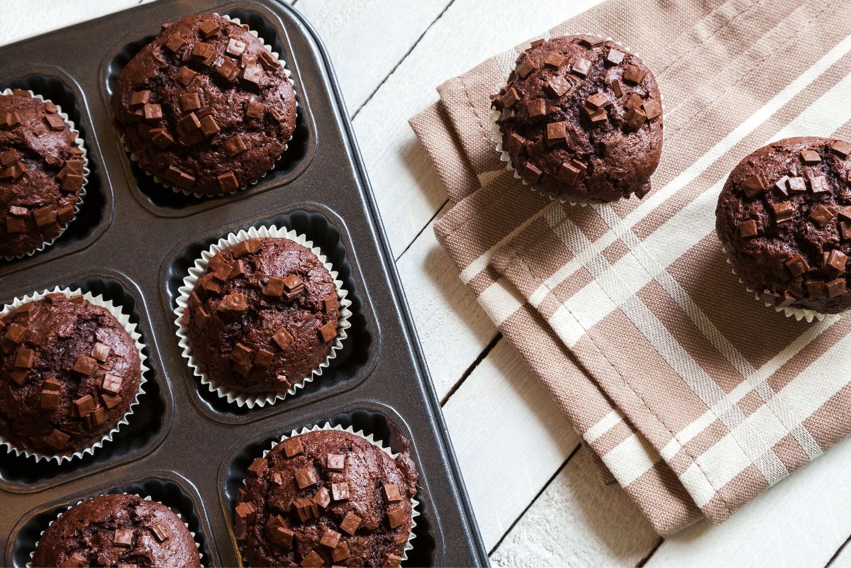 Chocolate oatmeal mini muffins topped with chocolate chunks, arranged in a dark muffin pan and on a folded brown plaid napkin over a white wooden table.