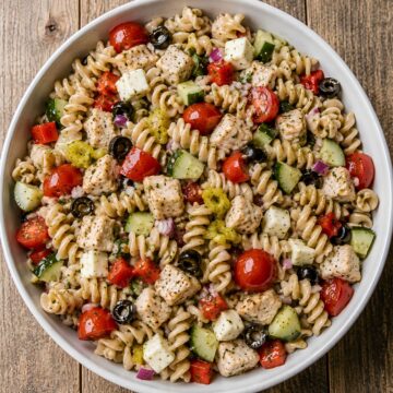 Overhead view of zesty Italian chicken pasta salad with whole wheat rotini, chicken, cherry tomatoes, cucumber, red bell pepper, red onion, pepperoncini, black olives, and mozzarella in a round white bowl on a rustic wood table.