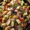 Close-up of zesty Italian chicken pasta salad with rotini, diced chicken, cucumbers, cherry tomatoes, red onion, black olives, and banana peppers in a black bowl.