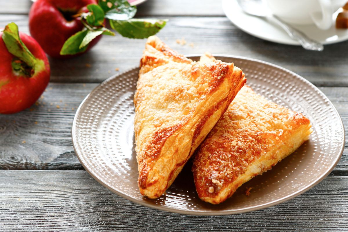 Two golden-brown air fryer apple turnovers on a plate, served on a rustic wood table with fresh red apples in the background.