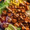 BBQ chickpea sweet potato bowl with roasted chickpeas and sweet potatoes, quinoa, corn, romaine lettuce, cherry tomatoes, red onion, sliced avocado, and creamy BBQ yogurt drizzle in a black bowl.