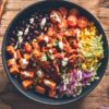Overhead view of a BBQ pulled chicken sweet potato bowl with roasted sweet potatoes, black beans, corn, cherry tomatoes, shredded cabbage, cilantro, and creamy dressing.