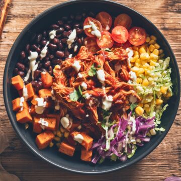 Overhead view of a BBQ pulled chicken sweet potato bowl with roasted sweet potatoes, black beans, corn, cherry tomatoes, shredded cabbage, cilantro, and creamy dressing.