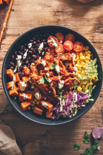 Overhead view of a BBQ pulled chicken sweet potato bowl with roasted sweet potatoes, black beans, corn, cherry tomatoes, shredded cabbage, cilantro, and creamy dressing.
