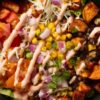 Close-up of black bean taco sweet potato bowl with roasted sweet potatoes, black beans, corn, tomatoes, lettuce, red onion, cheese, and creamy dressing.