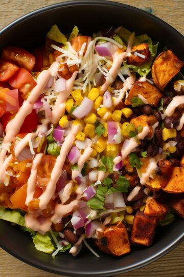 Close-up of black bean taco sweet potato bowl with roasted sweet potatoes, black beans, corn, tomatoes, lettuce, red onion, cheese, and creamy dressing.