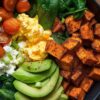 Close-up breakfast sweet potato protein bowl with roasted sweet potatoes, scrambled eggs, cottage cheese, spinach, cherry tomatoes, avocado slices, and green onions in a black bowl.