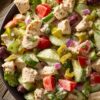 Chicken grinder chopped salad with diced chicken, romaine lettuce, cucumber, tomatoes, red onion, banana peppers, olives, and creamy dressing in a black bowl on a rustic wood table.