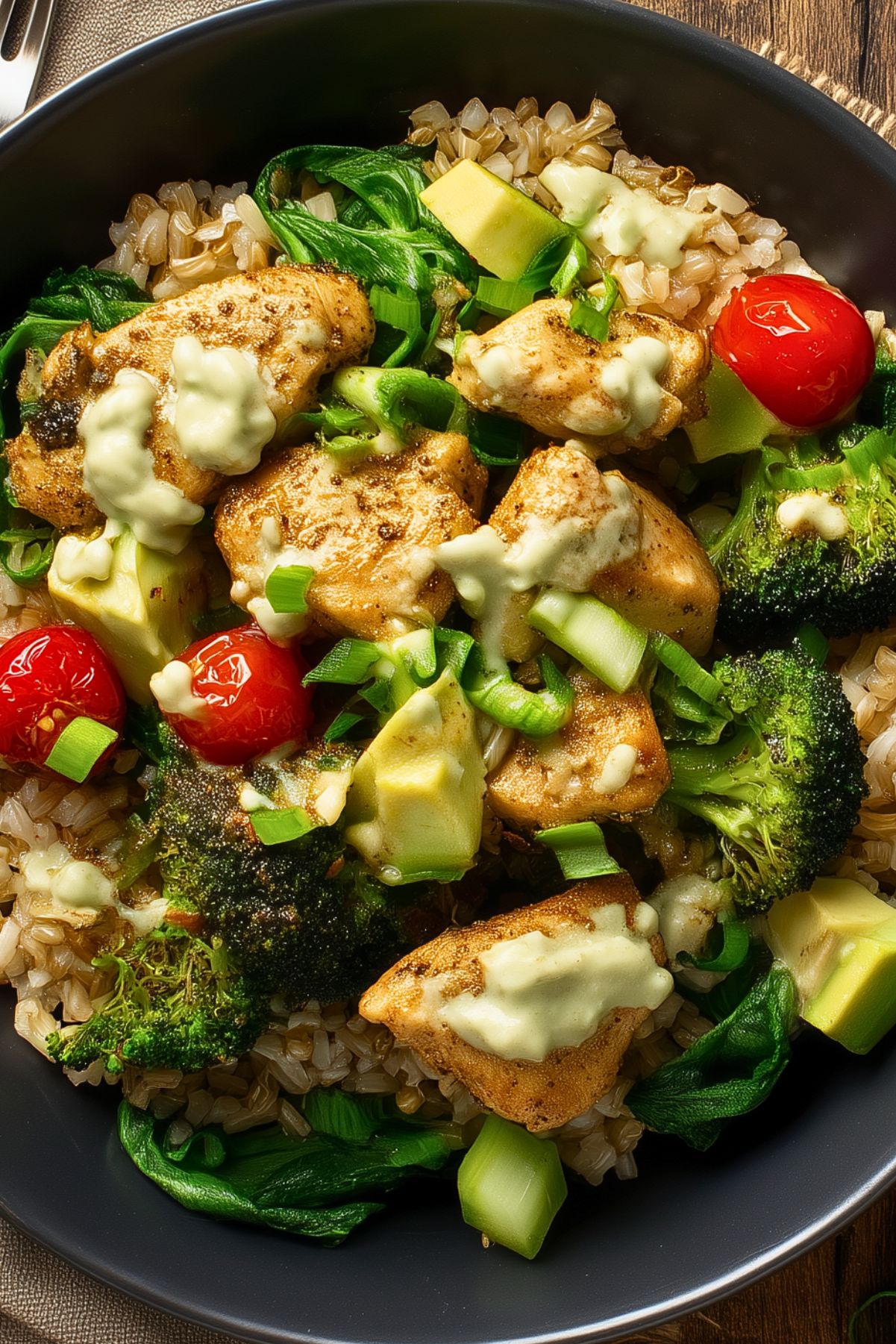 High protein chicken rice bowl with seasoned chicken breast, brown rice, broccoli, spinach, avocado, cherry tomatoes, green onions, and creamy dressing in a black bowl.