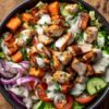 Overhead chicken shawarma sweet potato bowl with roasted chicken, sweet potato cubes, cucumber, tomatoes, red onion, lettuce, fresh herbs, and creamy sauce in a black bowl on rustic wood.