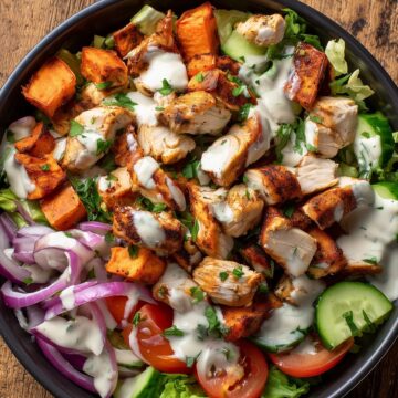 Overhead chicken shawarma sweet potato bowl with roasted chicken, sweet potato cubes, cucumber, tomatoes, red onion, lettuce, fresh herbs, and creamy sauce in a black bowl on rustic wood.