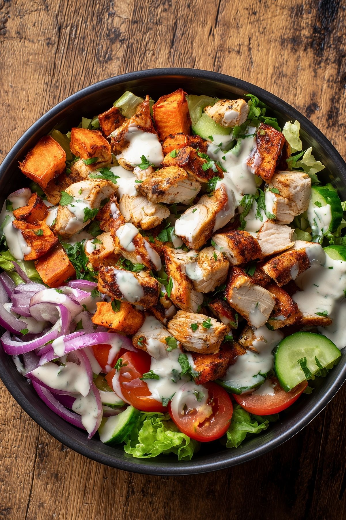 Overhead chicken shawarma sweet potato bowl with roasted chicken, sweet potato cubes, cucumber, tomatoes, red onion, lettuce, fresh herbs, and creamy sauce in a black bowl on rustic wood.