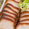 Sliced Chinese char siu pork loin with caramelized honey hoisin glaze arranged on a white platter and topped with curled green onions.