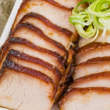 Sliced Chinese char siu pork loin with caramelized honey hoisin glaze arranged on a white platter and topped with curled green onions.