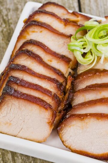 Sliced Chinese char siu pork loin with caramelized honey hoisin glaze arranged on a white platter and topped with curled green onions.