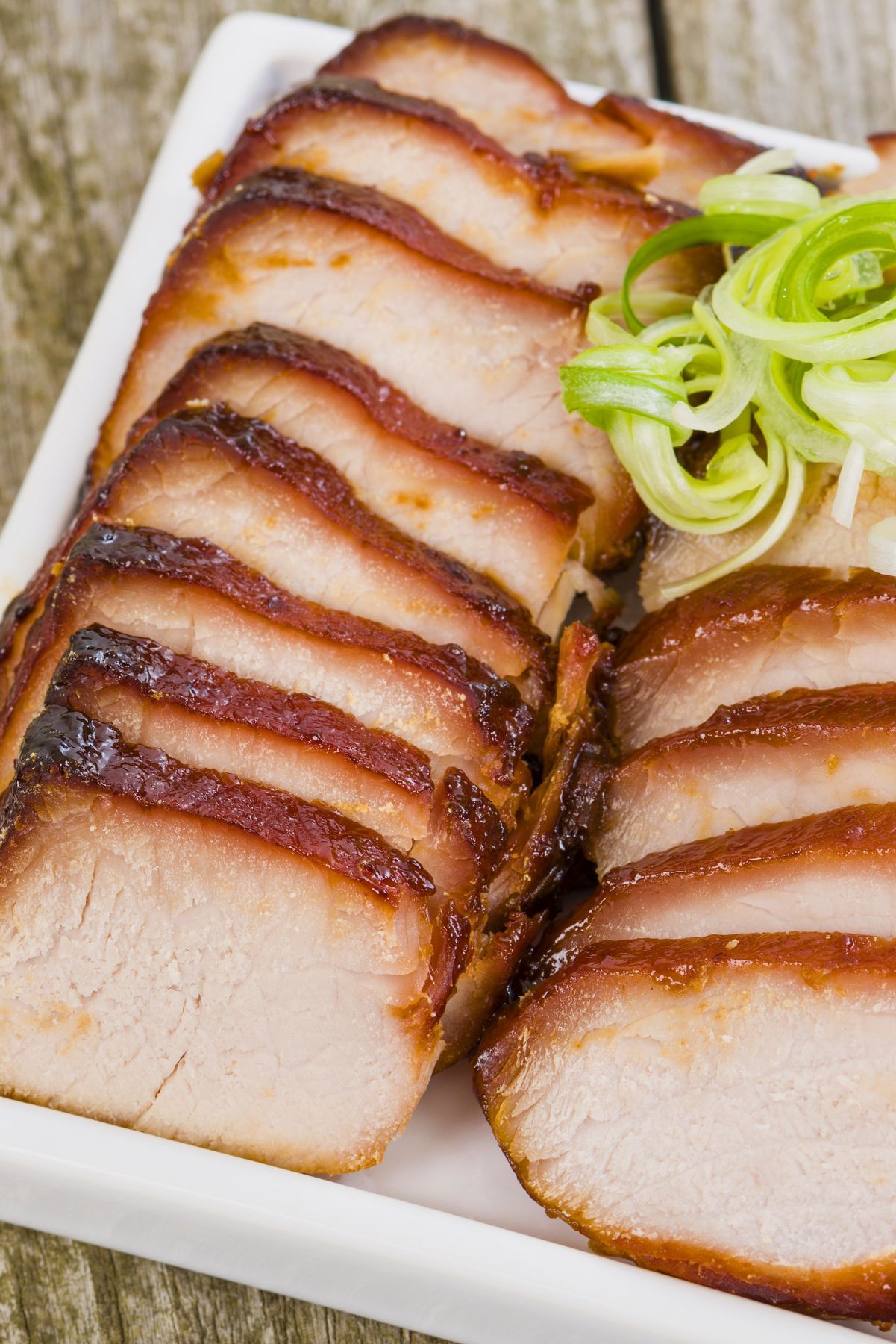 Sliced Chinese char siu pork loin with caramelized honey hoisin glaze arranged on a white platter and topped with curled green onions.