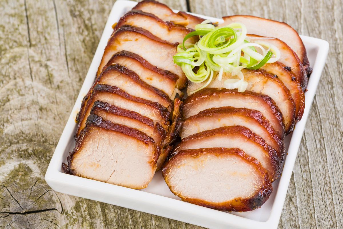 Chinese char siu pork loin sliced into thick pieces with a caramelized honey hoisin glaze, arranged on a white rectangular platter with curled green onion garnish on a rustic wood table.