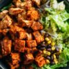 Chipotle chicken sweet potato bowl with seasoned chicken, roasted sweet potato cubes, black beans, corn, and crisp lettuce in a black bowl on a rustic wood table.