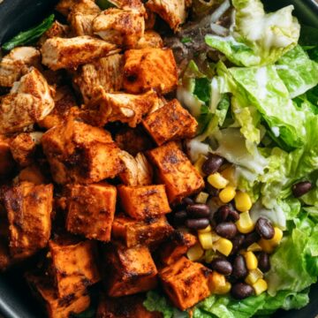 Chipotle chicken sweet potato bowl with seasoned chicken, roasted sweet potato cubes, black beans, corn, and crisp lettuce in a black bowl on a rustic wood table.