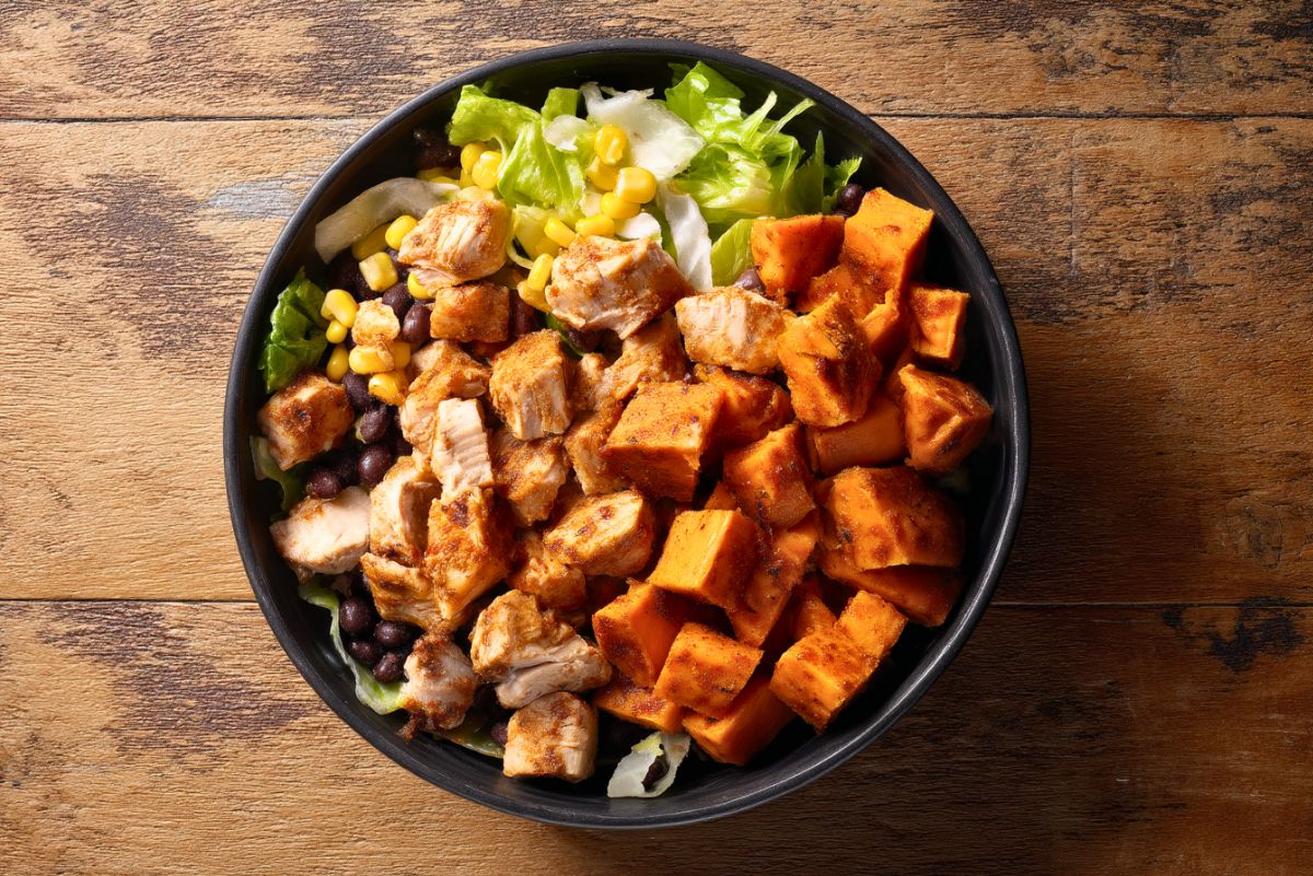 Chipotle chicken sweet potato bowl with seasoned chicken, roasted sweet potatoes, black beans, corn, and lettuce in a black bowl on a rustic wooden table.